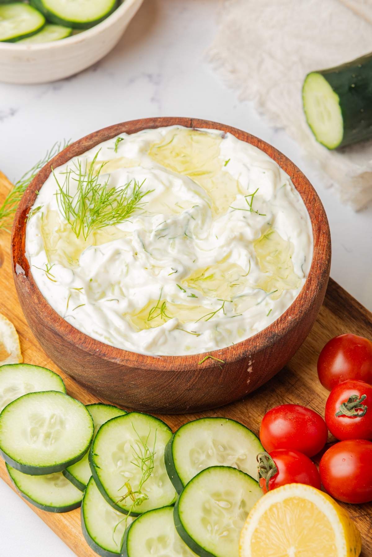 A wooden bowl filled with homemade tzatziki sauce, garnished with dill and surrounded by sliced cucumbers, cherry tomatoes, and a lemon wedge on a wooden board.