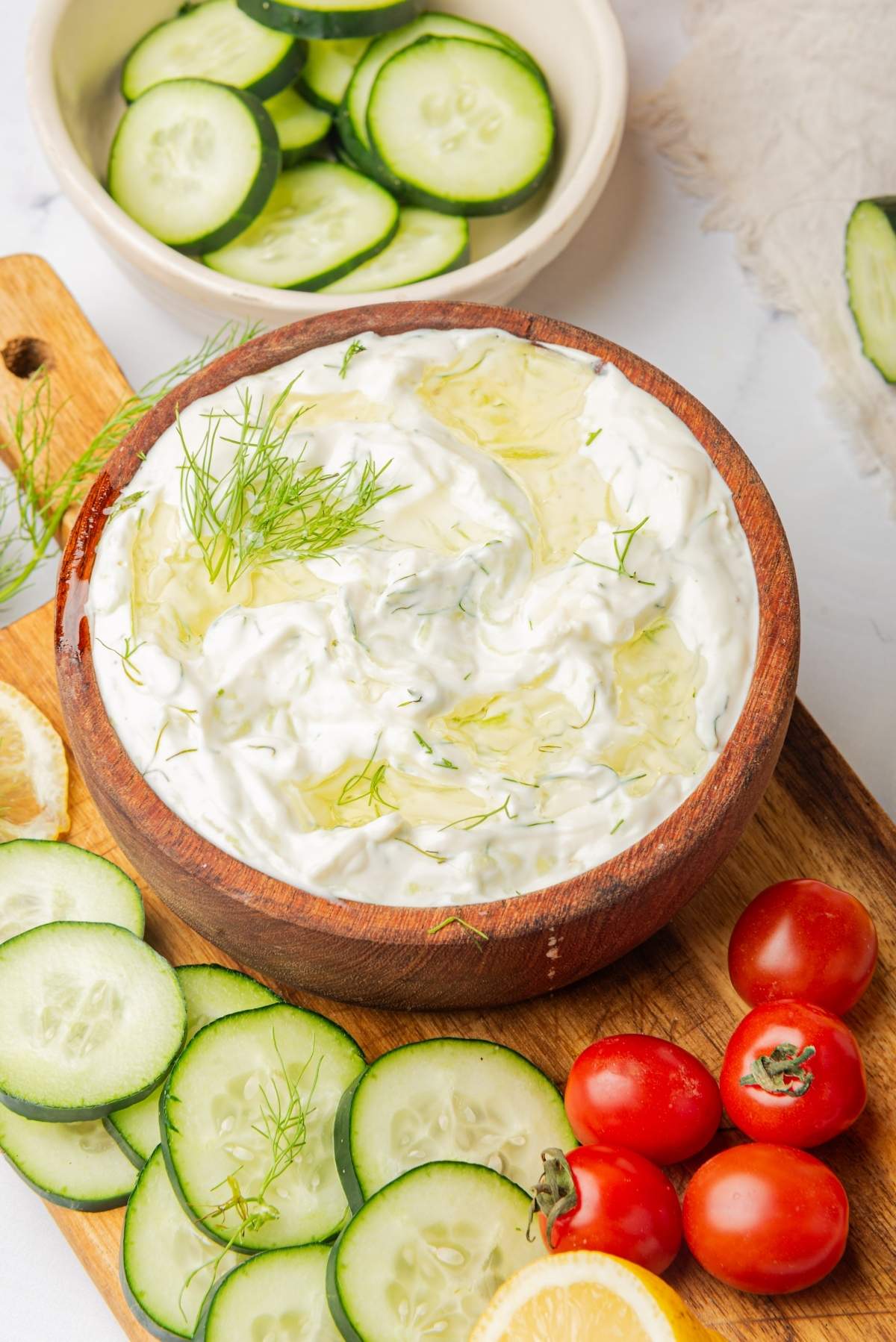 A wooden bowl filled with Greek-style tzatziki dip garnished with dill, surrounded by cucumber slices, cherry tomatoes, and lemon wedges on a wooden cutting board. A bowl of sliced cucumbers is in the background.