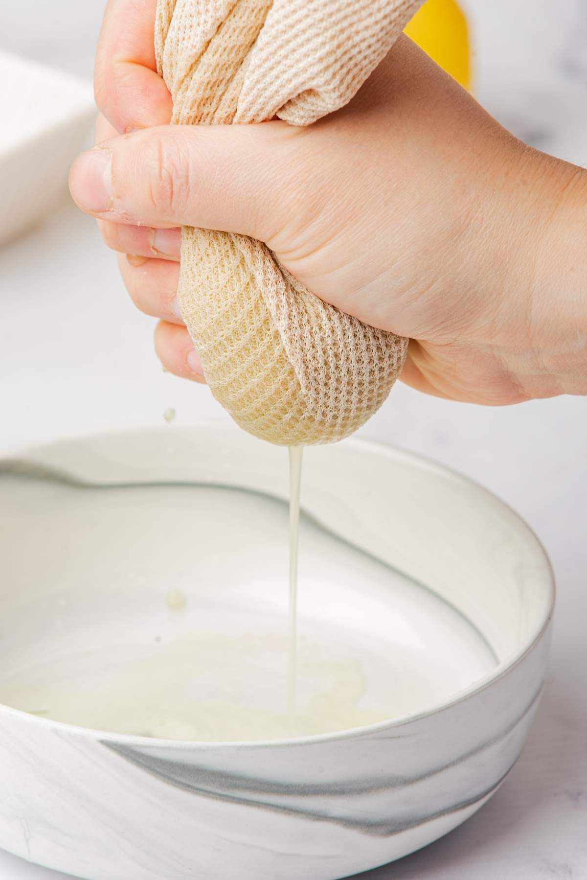 A hand is squeezing liquid from a beige cloth or towel into a white bowl, an essential step in preparing the perfect tzatziki recipe.