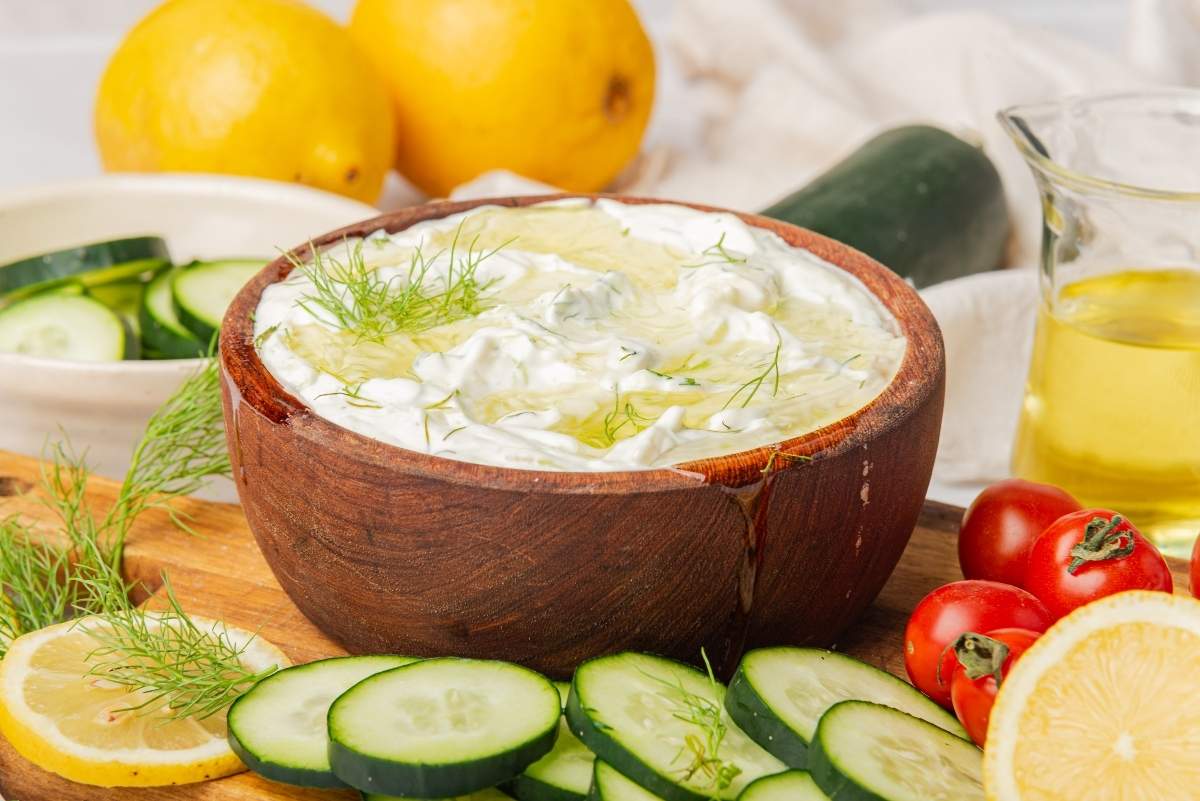 A wooden bowl filled with creamy Greek tzatziki sauce is surrounded by cucumber slices, cherry tomatoes, a lemon slice, and fresh dill. Nearby are whole lemons and a jug of olive oil.