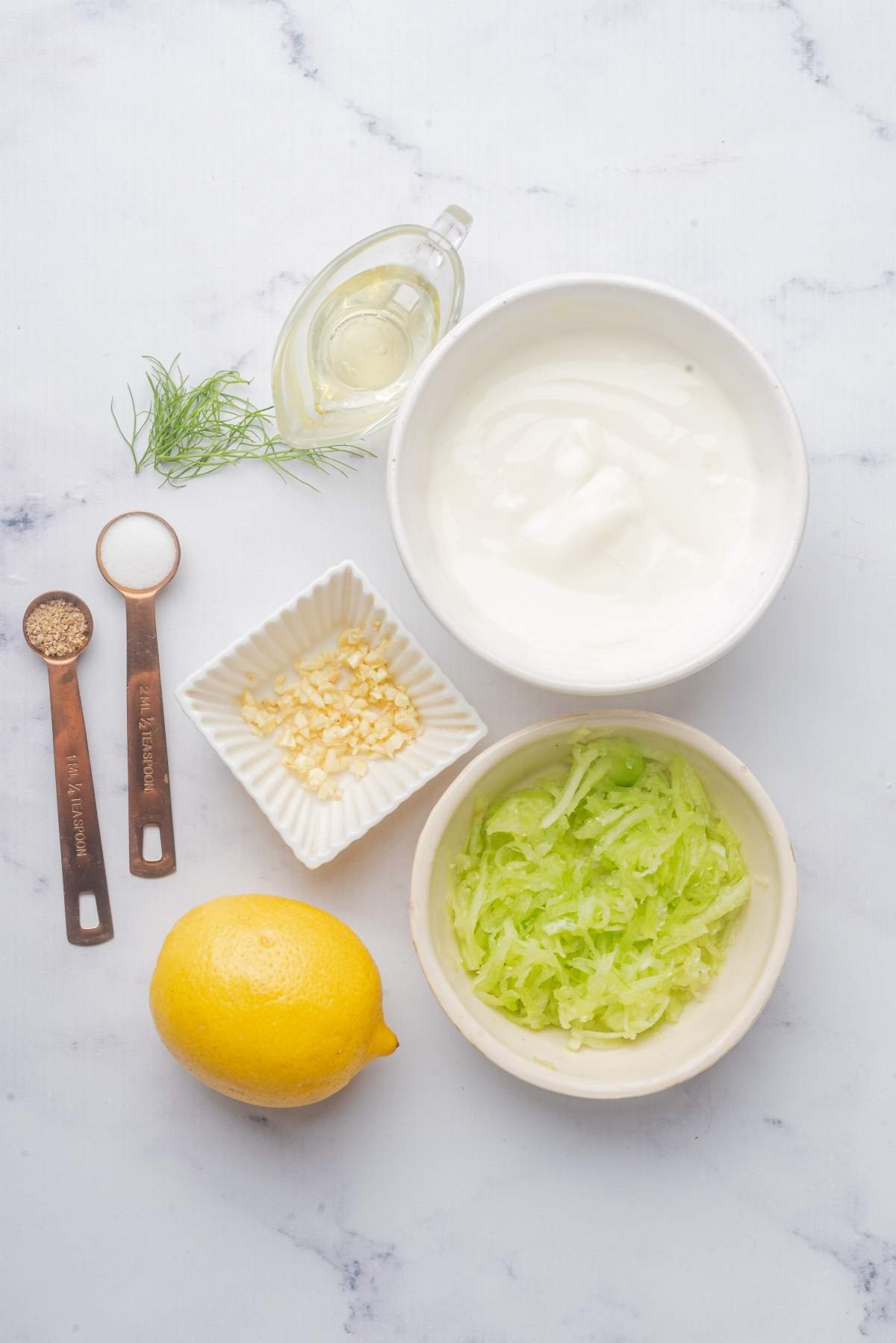 Ingredients for a delicious tzatziki recipe laid out: a bowl of yogurt, grated cucumber, chopped garlic, a lemon, olive oil, salt, pepper, and fresh dill on a marble surface.