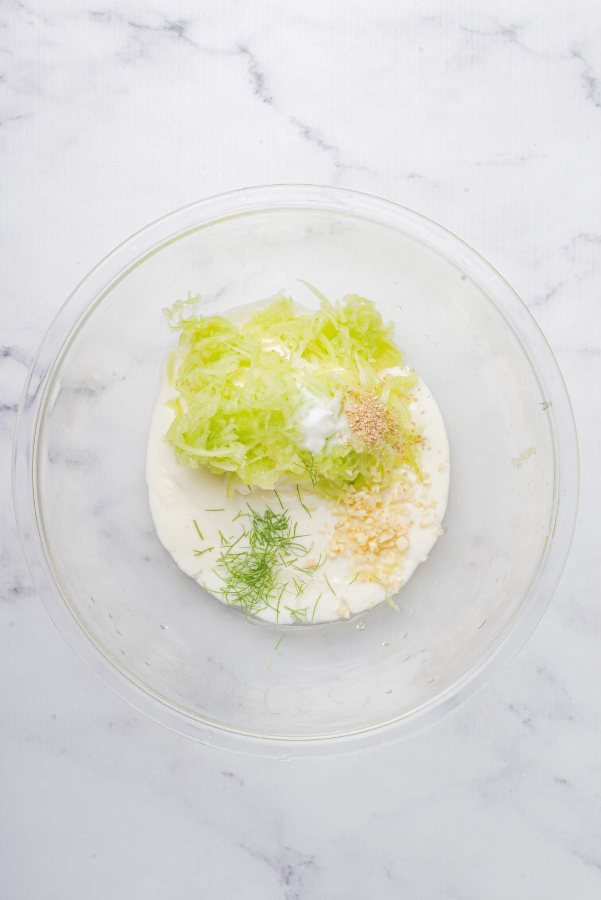 A clear glass bowl containing yogurt, shredded cucumber, minced garlic, dill, and a sprinkle of salt sits on a marble countertop—a classic tzatziki recipe.