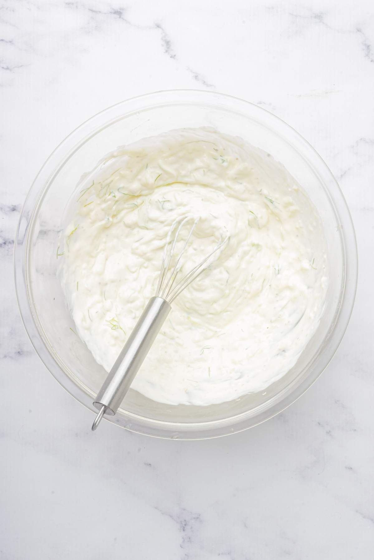 A mixing bowl filled with a creamy white mixture, reminiscent of a classic tzatziki recipe, with a whisk resting inside it on a white marble surface.