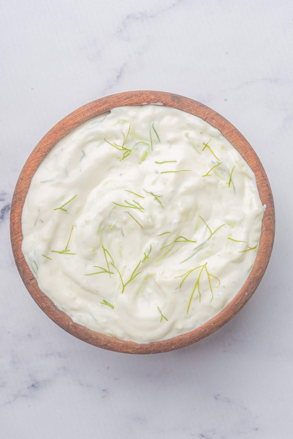 A wooden bowl filled with creamy white tzatziki sauce, garnished with thin slices of fresh dill, rests on a light marble surface, showcasing the perfect tzatziki recipe.