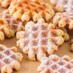 A batch of round, crispy waffle cookies, delicately sprinkled with powdered sugar, is arranged on a parchment paper-lined surface.