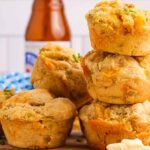 A stack of savory beer cheese muffins with visible herbs and ingredients, placed on a wooden surface. A bottle and kitchen cloth are in the blurred background.