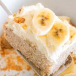 A slice of slow cooker banana cake topped with cream and banana slices being lifted from a dish.