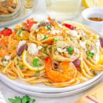 A plate of Mediterranean shrimp linguine topped with artichokes, olives, lemon slices, capers, and feta cheese sits on a table next to a fork and napkin, with ingredients and a wine glass in the background.