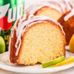 A slice of glazed 7 Up pound cake on a plate, surrounded by whole lemons and limes.