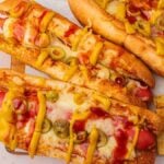 Air fryer pizza dogs topped with melted cheese, sliced olives, mustard, and ketchup are served with a side of fries on a wooden board.