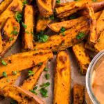 Air fryer sweet potato wedges, garnished with herbs, are served on parchment paper alongside a small bowl of creamy dipping sauce.