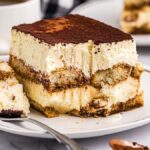 A slice of authentic Italian tiramisu rests on a white plate with a spoon beside it. Coffee cups are visible in the background, highlighting the perfect pairing for this classic treat.