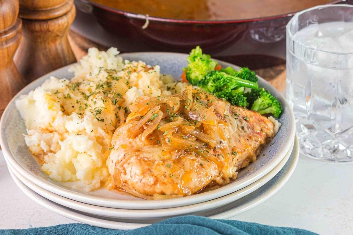 Plate with creamy mashed potatoes and French onion chicken, seasoned to perfection with caramelized onions, accompanied by a side of steamed broccoli. A glass of ice water sits nearby.