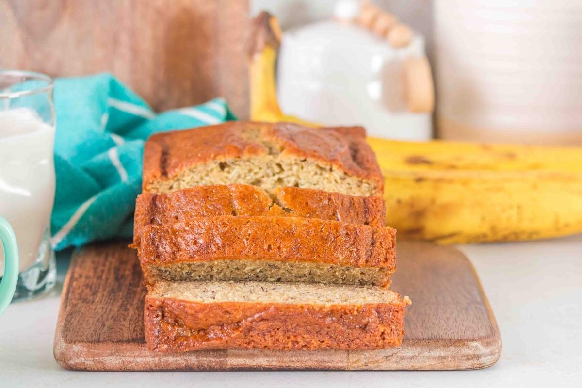 A sliced loaf of brown butter banana bread sits on a wooden cutting board, with a ripe banana and a blue towel in the background.