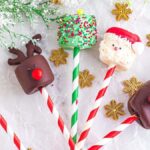 Christmas marshmallow pops decorated as reindeer and Santa, with sprinkles, on red and white striped sticks. Surrounded by snowflake decorations and greenery on a marble background.
