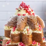 A holiday-themed pandoro Christmas tree cake with layers of pastry and cream, topped with powdered sugar and pomegranate seeds. Resembling a Christmas tree, it's festively decorated with greenery in the background.