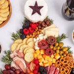 A festive Christmas tree charcuterie board arranged with cheese, meats, berries, grapes, nuts, crackers, pretzels, and a glass of red wine. Nearby sits a bowl of jam and dried apricots to complete the cozy holiday spread.