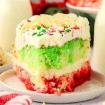 A slice of festive Christmas poke cake with vibrant red, green, and white layers sits invitingly on a plate, topped with cheerful sprinkles. In the background, two glass bottles filled with milk complete the delightful holiday scene.