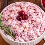 A bowl of pink cranberry fluff salad topped with a few whole cranberries, chopped nuts, and a sprig of rosemary sits next to a spoon.