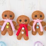 Five felt gingerbread man ornaments with colorful scarves and buttons are lined up on a white surface, surrounded by scattered festive decorations.