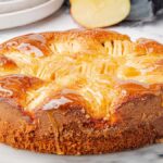 A glazed apple ginger cake rests on a marble surface, surrounded by whole and sliced apples.