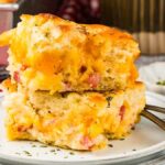 Two slices of ham and cheese butter swim biscuits on a plate, garnished with herbs. The scene evokes the comfort of ham and cheese butter swim biscuits. Silver forks rest beside the plate, ready to dive into this savory delight.