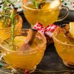 Four glass cups filled with a spiced holiday punch, garnished with cinnamon sticks, apple slices, and rosemary, are arranged on a tray adorned with decorative beads.