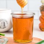 A jar of jalapeño hot honey with a honey dipper above it sits enticingly on a wooden board. Nearby, a bear-shaped honey container and fresh jalapeños add a spicy twist to the scene.