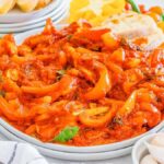 A plate of peperonata, rich with red pepper stew, sits alongside crusty bread. It's surrounded by vibrant whole peppers, garlic cloves, and a bottle of olive oil.