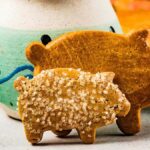 A pair of delightful marranitos cookies, one adorned with sugar sprinkles, sit charmingly in front of a decorative ceramic jug.
