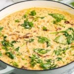 A pot filled with creamy Tuscan marry me chickpeas, garnished with green spinach leaves and herbs, sits on a white surface.