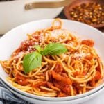 A bowl of classic pasta amatriciana, topped with grated cheese. A blue patterned napkin complements the scene, surrounded by bowls of red pepper flakes and parmesan.