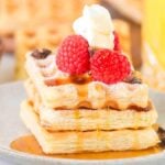A stack of puff pastry waffles topped with raspberries, whipped cream, chocolate chips, and maple syrup sits invitingly on a plate.