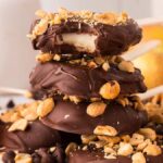 A stack of chocolate-covered apple slices with peanuts sits invitingly, surrounded by fresh apples, reminiscent of delightful Chocolate Caramel Apple Pops.