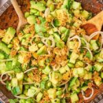 A wooden bowl brimming with a crispy rice salad, featuring diced cucumber, edamame, and crispy onions. It's surrounded by small bowls of herbs and spices, with two wooden spoons artfully placed for serving.