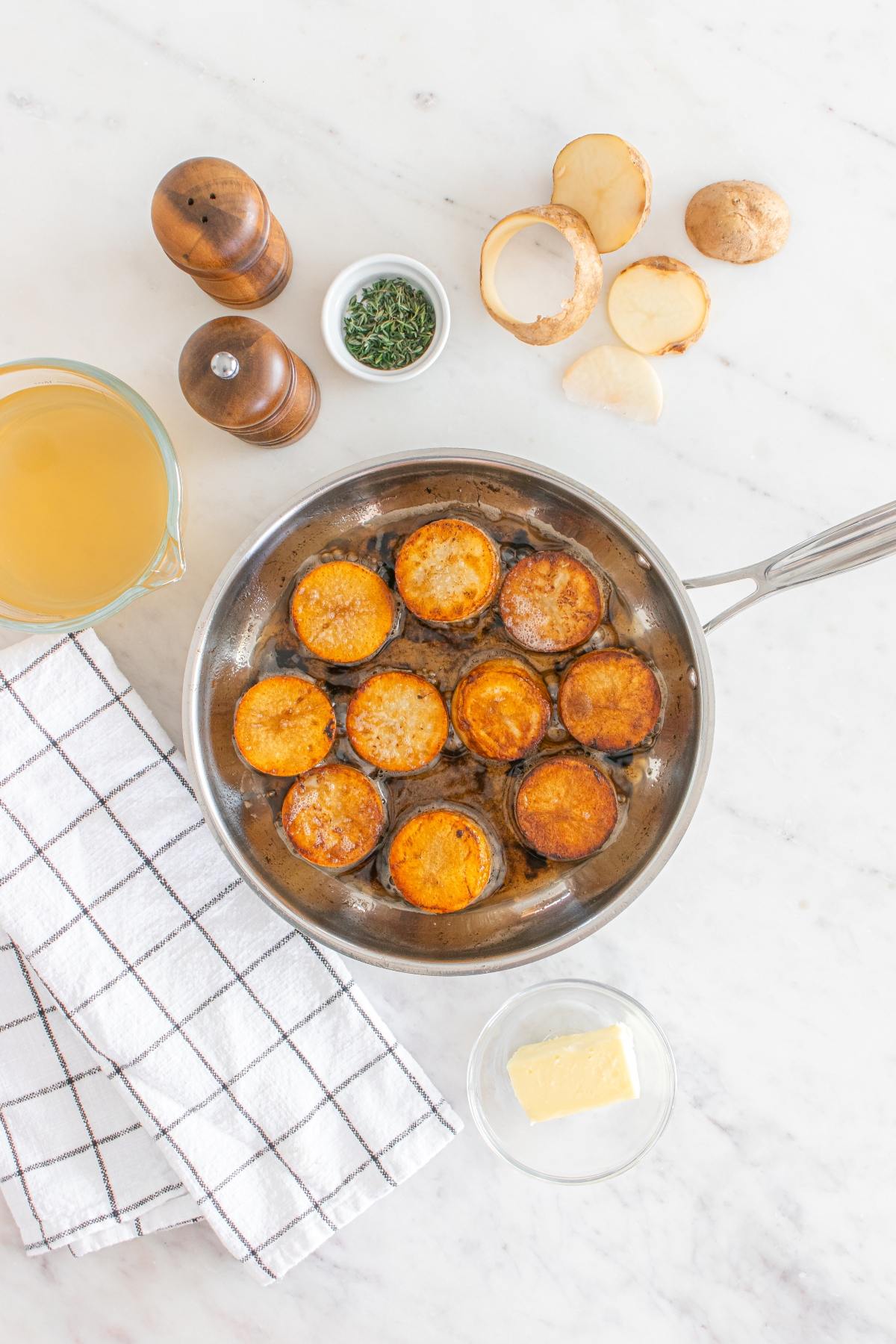Sliced potatoes browning in a pan, with garlic butter fondant potatoes, broth, and fresh herbs arranged on a marble countertop.