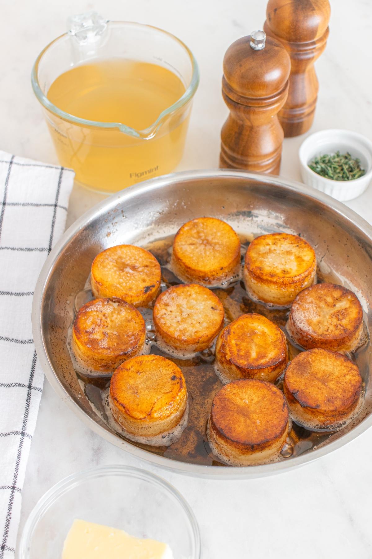 Round, golden-brown garlic butter fondant potatoes cook in a skillet with broth, herbs, salt, and pepper for a rich and savory side.