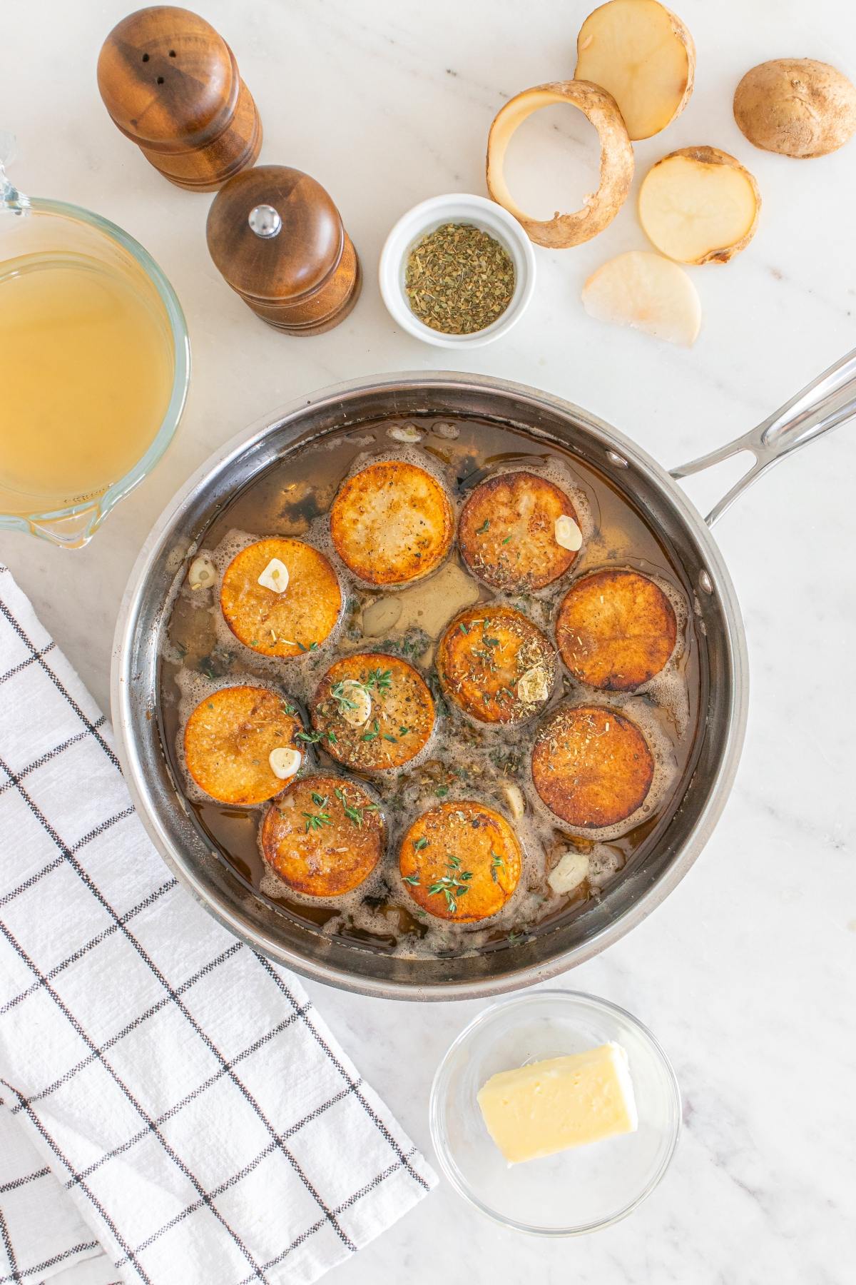 Garlic butter fondant potatoes simmer as sliced potatoes infuse with garlic, herbs, broth, and butter on a marble countertop.