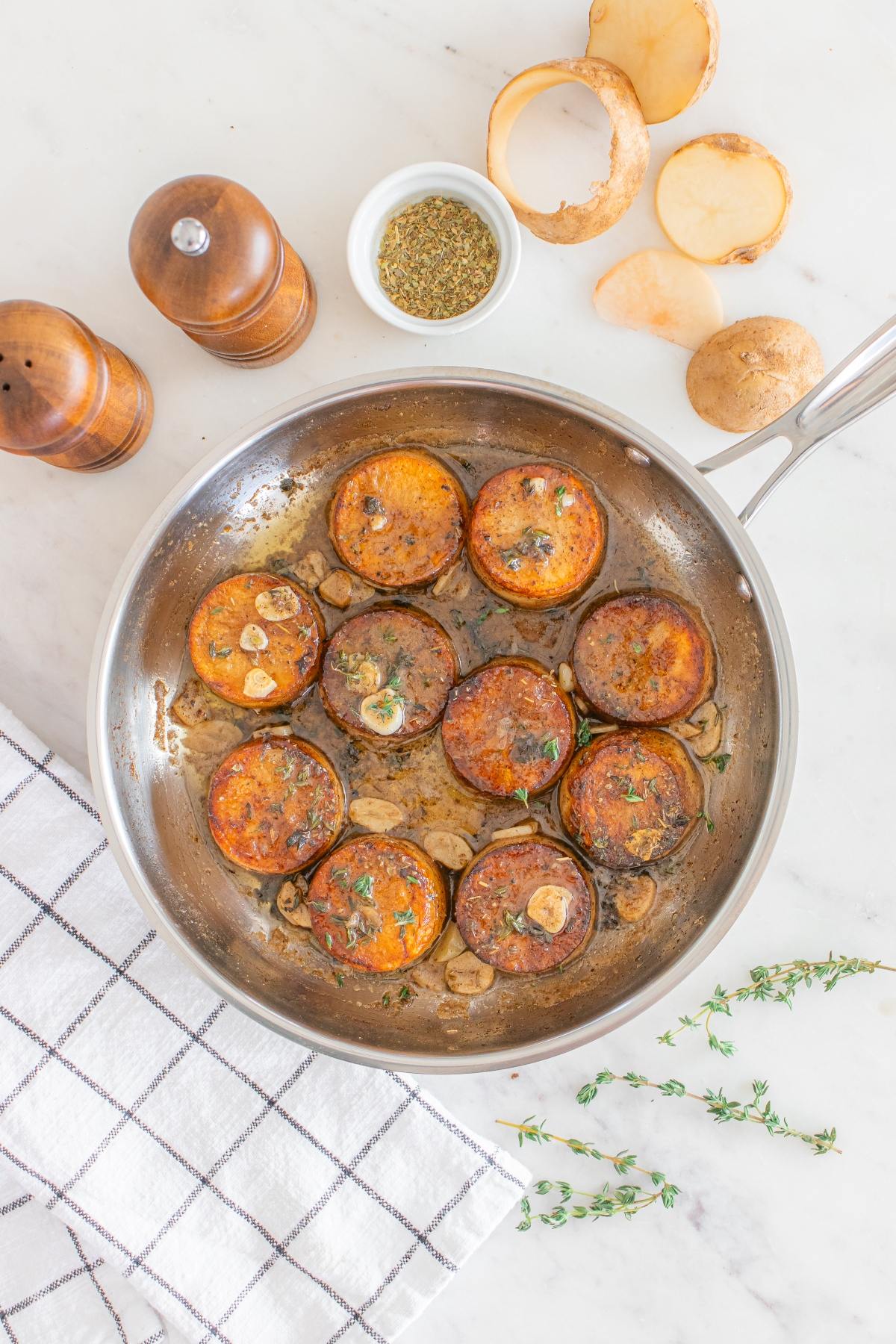 Sliced potatoes saut&eacute;ing in a pan with herbs and garlic, surrounded by seasonings and fresh potatoes.