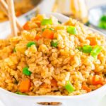 A bowl of hibachi fried rice with diced carrots and green onions, garnished with additional green onions on top. Chopsticks are artfully placed in the bowl.