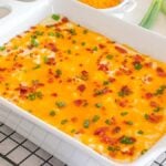 A casserole dish filled with a cheesy, bacon-topped loaded mashed potato casserole sprinkled with green onions rests on a cooling rack.