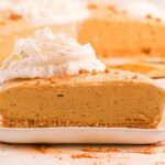 A slice of no bake gingerbread cheesecake with whipped cream sits on a white plate, surrounded by crumbs and a fork nearby.