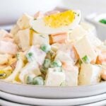 A plate of authentic Olivier salad features diced potatoes, carrots, peas, and a sliced boiled egg on top, all garnished with fresh dill. In the background, a small bowl of green peas complements this classic Russian dish perfectly.