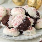 A plate of peppermint mocha cookies partially dipped in white frosting, sprinkled with crushed peppermint candies, surrounded by red and white mints and festive decorations.