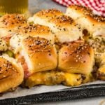 A tray of bacon cheeseburger sliders with sesame seed buns, melted cheese, crispy bacon, and slices of pickles. A red and white checkered cloth and two glasses of beer in the background add to the cozy setting.