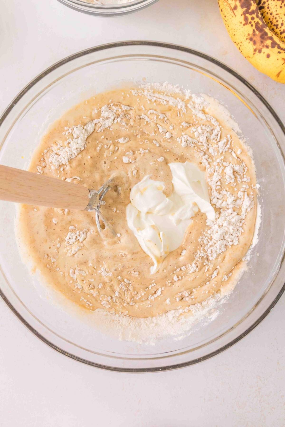 Mixing bowl with brown butter banana bread batter, flour, and a dollop of cream on top. Wooden spatula resting in the batter. Bananas visible in the corner.