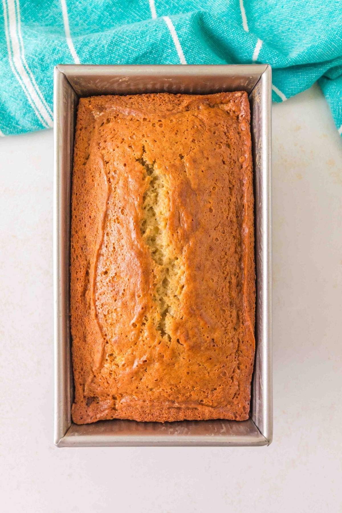 A loaf of brown butter banana bread rests in a rectangular metal pan, set on a light-colored surface with a turquoise cloth gently draped nearby.