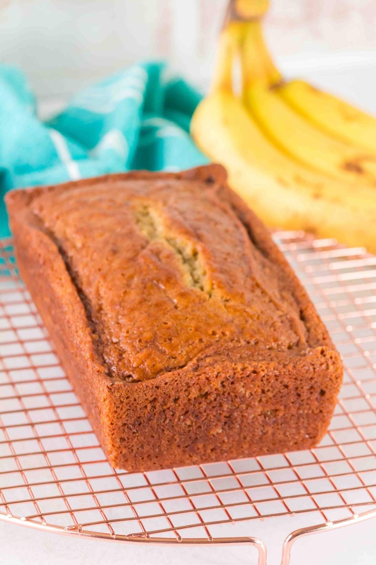 A loaf of brown butter banana bread rests on a cooling rack, surrounded by ripe bananas and a blue cloth in the background.