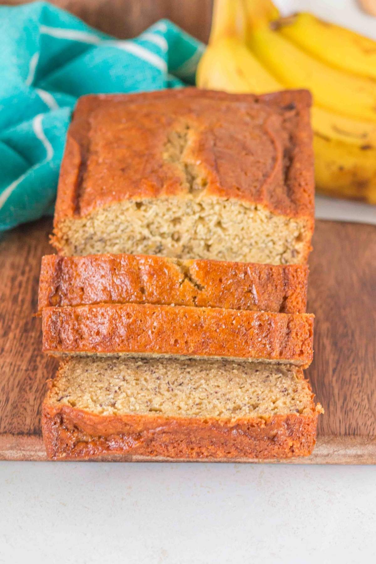 A sliced loaf of brown butter banana bread rests on a wooden board, surrounded by a bunch of bananas and a blue cloth in the background.