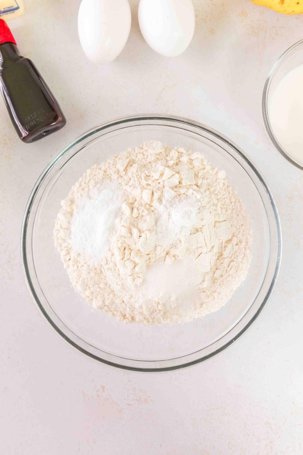 A glass bowl with dry baking ingredients sits on the countertop, ready to create a delicious brown butter banana bread. It's surrounded by vanilla extract, eggs, and a bowl of milk, all waiting to work their magic together.